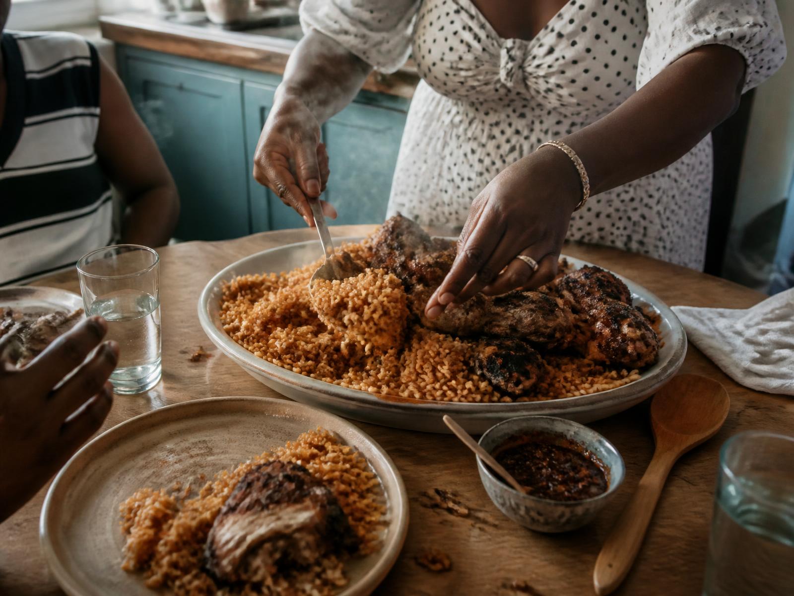 Hands serving a big platter of jollof rice and grilled chicken onto plates at a wooden family table
